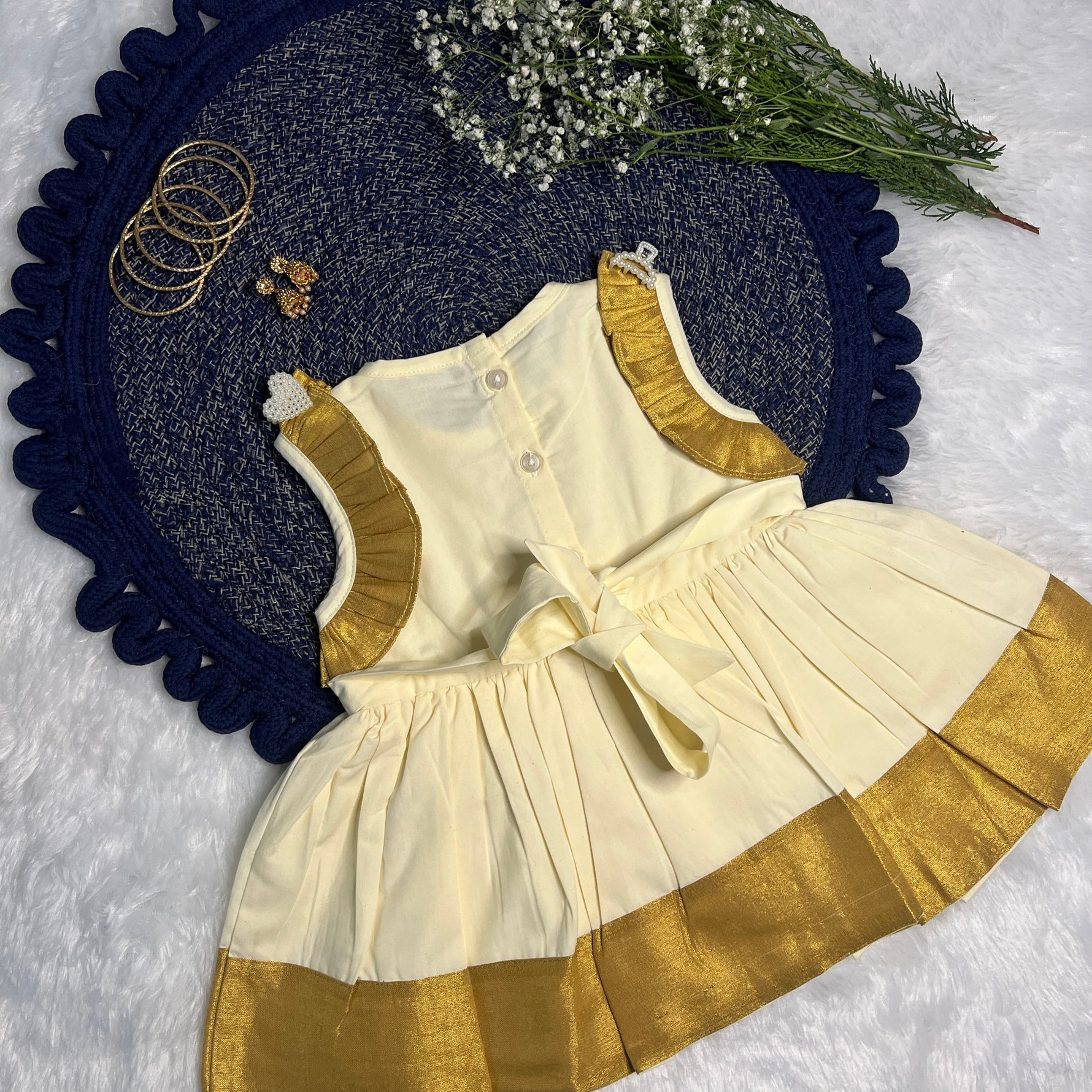Children's outfit with white top and gold trim on a textured surface with flowers and jewelry.