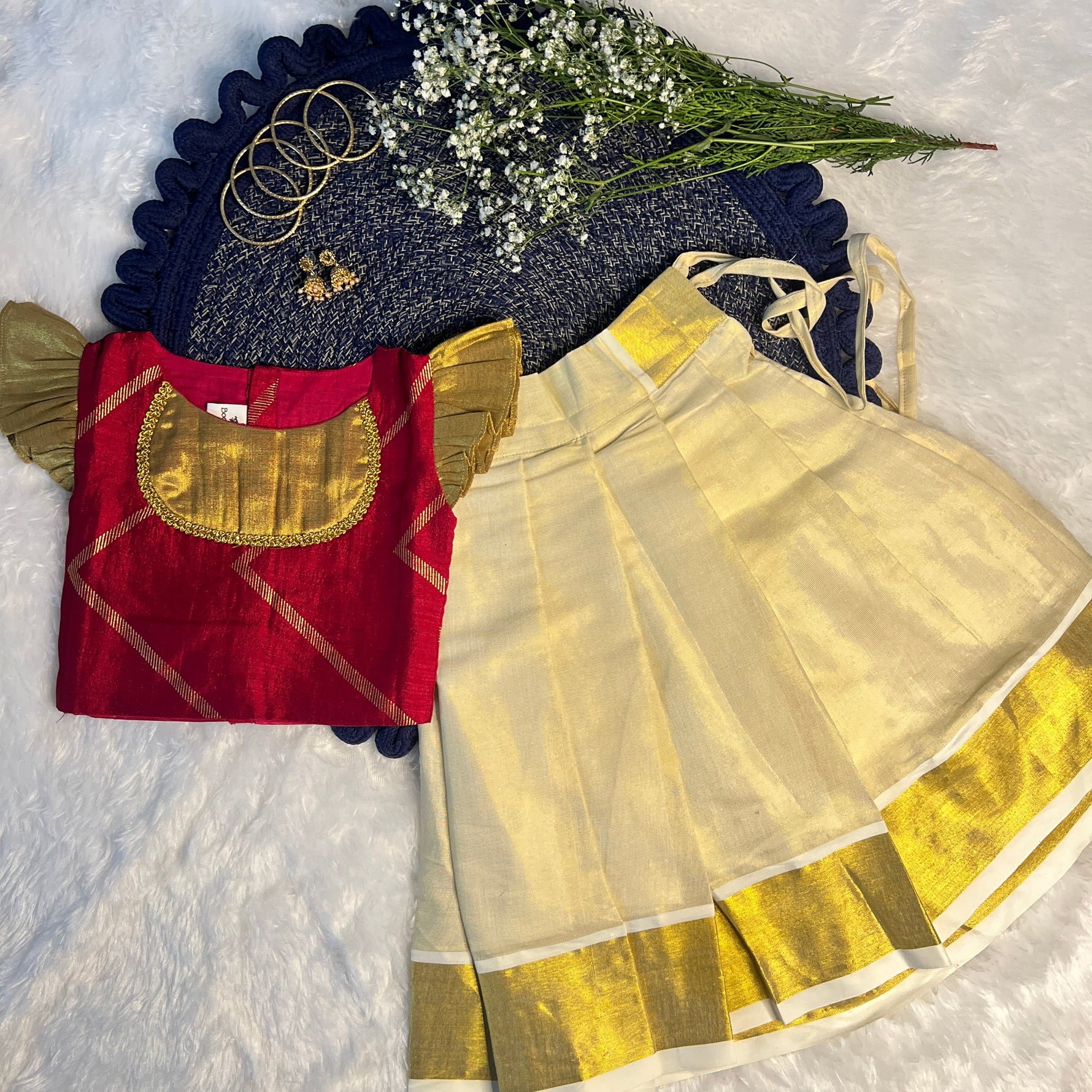 Traditional red and gold blouse with a white saree with gold border on a textured surface.