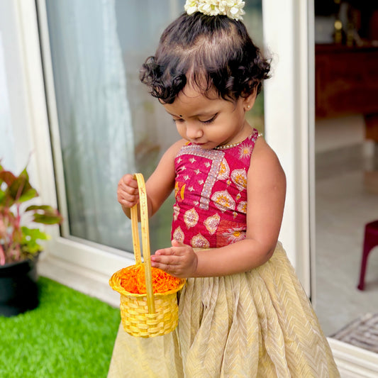 Child in an ethnic dress, red top and yellow skirt holding a basket outdoors.